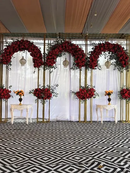 A bold and dramatic outdoor stage backdrop featuring arches of red roses against a black and white geometric floor. The design is modern, striking, and perfect for a reception.