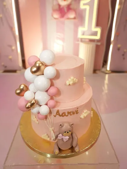 A sweet pink teddy bear cake with a balloon garland decoration.