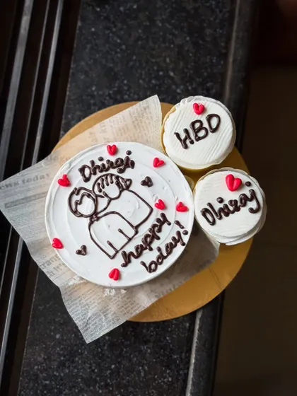 This is another view of the personalized birthday set for Dhiraj, showing the bento cake and cupcakes on a wooden board. The hand-drawn style gives it a really personal feel.