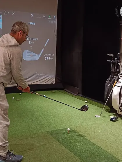 A client prepares to hit a shot in the simulator, with the club's dynamic lie and impact height data displayed on the screen for analysis.