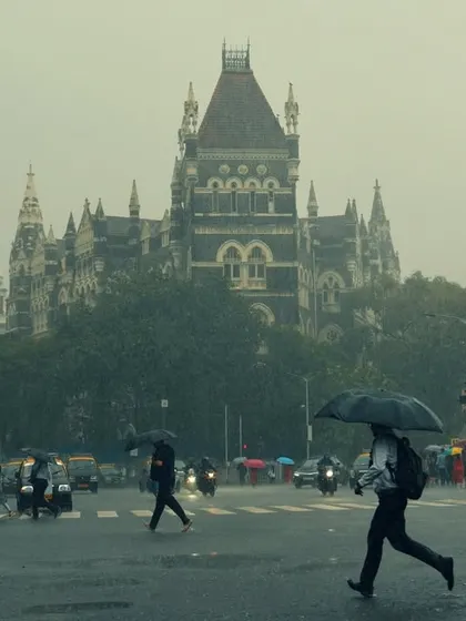 Pedestrians cross a wet street with the historic High Court building looming in the background. The image captures the daily rhythm of Mumbai life that continues, beautifully and stoically, even in the middle of a downpour.