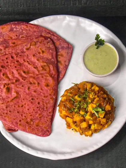 A unique Indian meal of beetroot paratha served with a savory paneer masala and mint chutney. The dark background makes the vibrant pink of the paratha stand out.