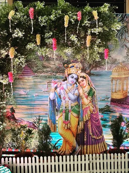 A beautiful photo booth with a Radha Krishna theme. The backdrop is a serene painting of the divine couple, framed by a real tree decorated with floral hangings.