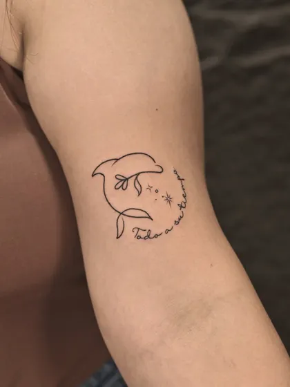 A playful and simple single-line dolphin tattoo. The design incorporates a flower and sparkling stars, with a delicate script that reads "Todo a su tiempo" (Everything in its own time).