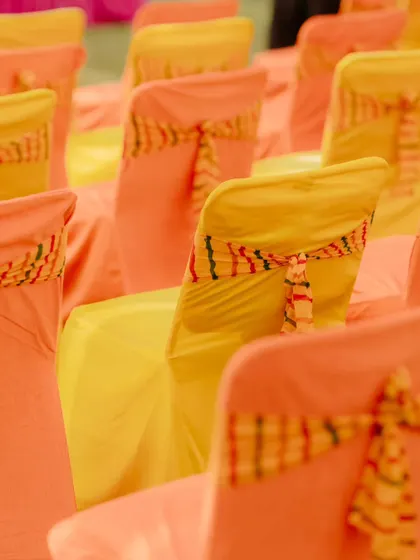 A close-up of the seating arrangement, showing chairs covered in yellow and orange fabric. The custom ties on the chair backs match the colourful theme of the Haldi celebration.