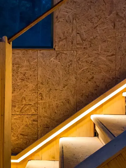 The staircase design incorporates integrated LED lighting along the handrail, providing safe and ambient illumination while highlighting the texture of the OSB wall panels.