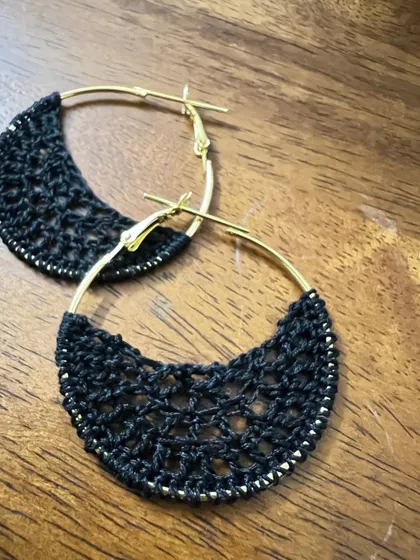 A close-up of the black half-moon hoop earrings. The delicate crochet work is edged with a fine golden thread.