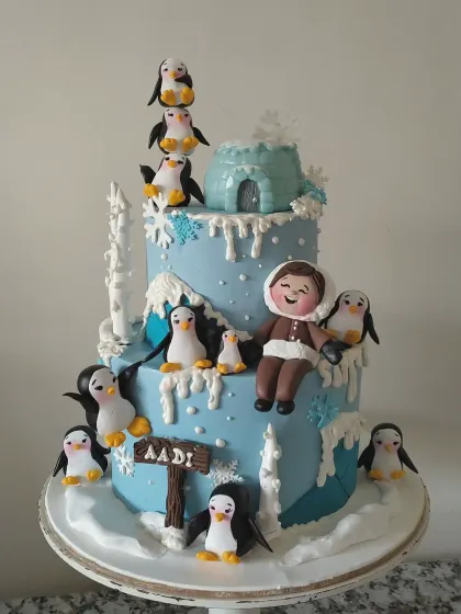 A winter wonderland adventure for a special birthday boy. This two-tiered cake is covered in snowy details, a fondant igloo, and a whole colony of adorable, hand-sculpted penguins. The personalized figurine of the birthday boy makes him the star of this chilly, charming scene.