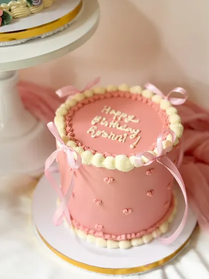 A sweet pink birthday cake with vintage-style piping. The design includes delicate heart imprints on the side and is finished with pretty pink ribbons.