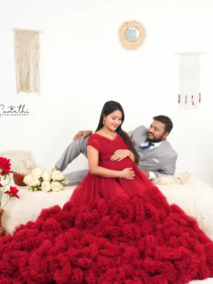A full-length view of the couple in a boho-chic setup. The wife's red gown contrasts beautifully with the neutral tones of the macrame and cushions.