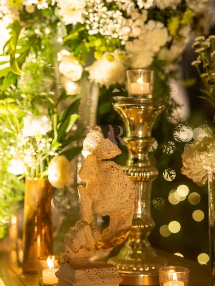 A close-up of a gold candelabra and a small sculpture, surrounded by white flowers and the soft bokeh of fairy lights. This is how I layer light and texture to create depth and romance.