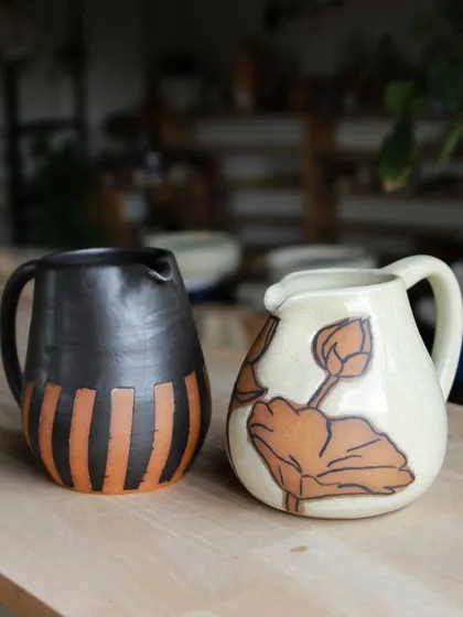 Two pitchers made by students, each with a unique surface decoration. One features a wax-resist pattern, while the other has a delicate, hand-painted lotus design.