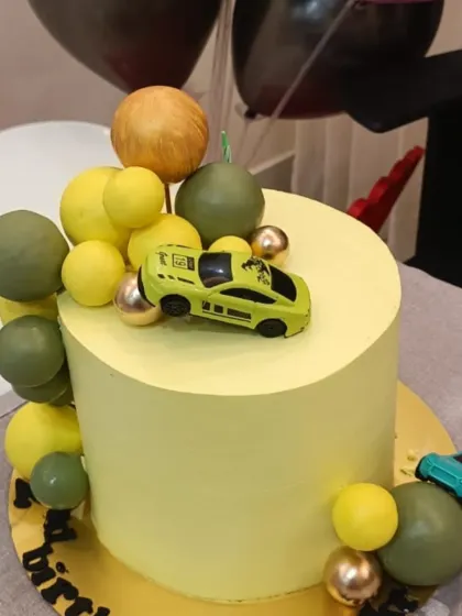 A modern car cake with a yellow and olive green color scheme. The cake is decorated with a toy car and a cascade of chocolate balls.