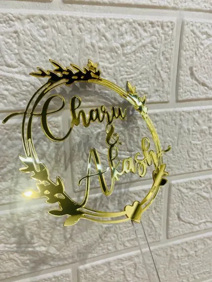 A close-up of the "Charu & Akash" wreath topper against a white brick background.
