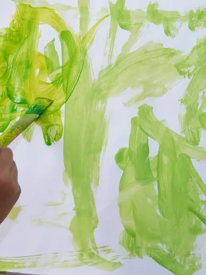 A close-up of a child's hand painting with bold green strokes. Art is a sensory experience, and we encourage children to explore the feel and flow of the paint as much as the final image.
