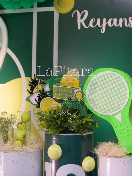 Detailed props on the tennis stage, including a giant tennis racket, balls, and lush green foliage.