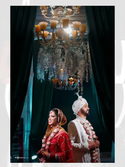 The bride and groom standing back-to-back under a magnificent chandelier after their wedding ceremony. This stylish portrait captures their stunning traditional attire and the opulence of the wedding decor.