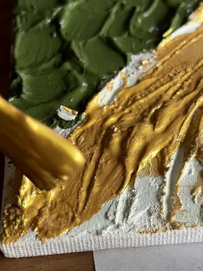 A close-up of a brush adding gold paint to a textured canvas. The combination of texture and shimmer creates a truly luxurious and dynamic artwork.