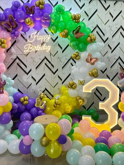 A vibrant unicorn and butterfly themed birthday party for a 3-year-old. The setup features a rainbow-colored balloon arch, a "Happy Birthday" neon sign, and a large light-up number "3".