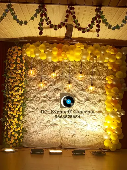 A simple yet elegant birthday backdrop. The design uses a textured white rose wall framed by yellow balloons and flowers, with hanging Edison bulbs adding a warm and celebratory glow.