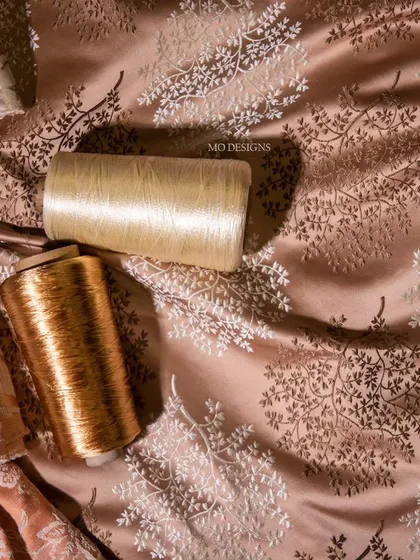 The raw materials of our craft. Spools of fine golden and cream thread lie on a lustrous fabric with delicate botanical embroidery, showcasing our dedication to handcrafted luxury.