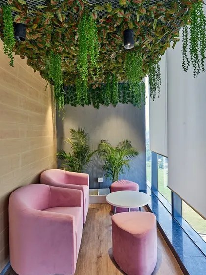 A small lounge area with pink chairs and a ceiling covered in greenery. Breakout spaces are essential for employee well-being. This cozy lounge corner, with its vibrant pink chairs and a ceiling canopy of artificial leaves, provides a refreshing escape from the main work floor.
