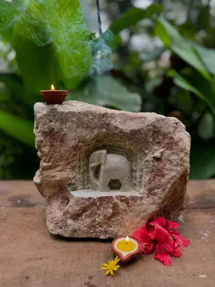 An elephant carved into a natural rock niche, with a diya and incense creating a devotional mood. The artist retains the stone's rustic originality while revealing a beautifully detailed character within.