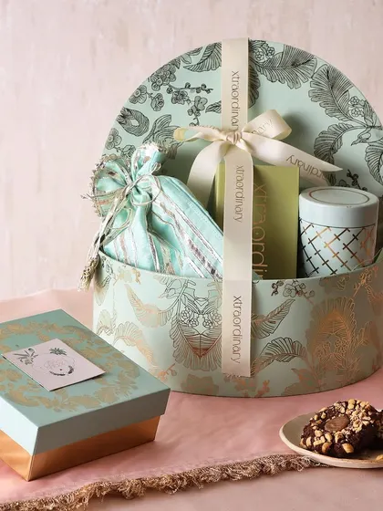 The Minted Joy hamper is a round, beautifully designed box filled with a slider box of hazelnut cookies, sugar-free mithai, honey choco almonds, and a tea cake or candle.