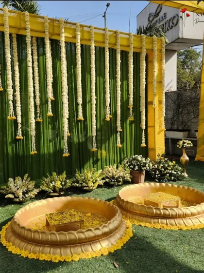 A simple yet beautiful Haldi setup. The backdrop of green drapes with hanging jasmine flowers provides a fresh and natural look, complemented by the traditional brass 'urlis'.