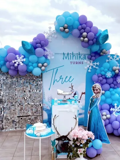 I created this winter wonderland for Mihika's third birthday with a Frozen theme. The rooftop setup includes character cutouts of Elsa and Olaf, a shimmering silver sequin wall, and a balloon arch in icy shades of blue and purple.