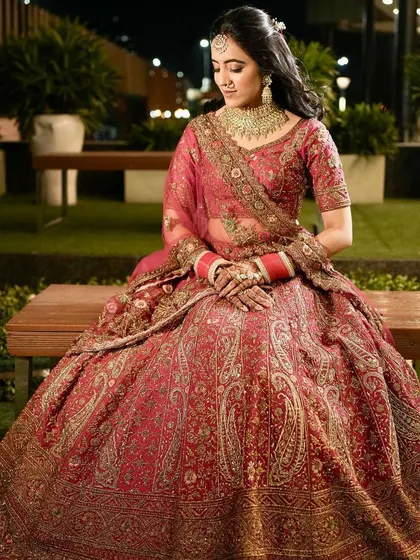 A full-length portrait of the bride seated in an outdoor setting at night. The lighting highlights the rich paisley patterns on her beautiful red lehenga.