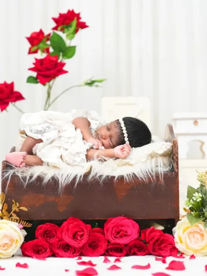 A sleeping newborn dressed in a delicate white lace outfit on a tiny bed surrounded by red and white roses. This elegant setup creates a timeless and classic portrait.