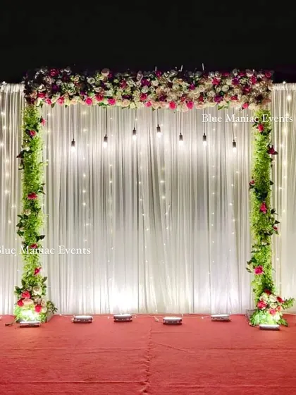 An elegant stage backdrop with a white fairy light curtain and pillars of greenery and flowers. The hanging Edison bulbs add a touch of industrial chic to the otherwise classic design.