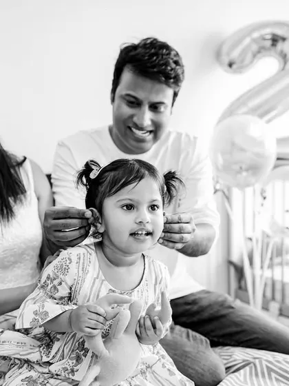 A candid black and white family photo from a second birthday shoot. The parents are sharing a laugh while their daughter looks on, capturing a genuine slice of their family life.