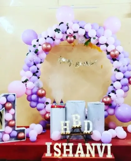A stylish first birthday decoration for Ishanvi on a raised stage. It features a circular balloon arch in purple and rose gold, a large "1" prop filled with balloons, and light-up name letters.