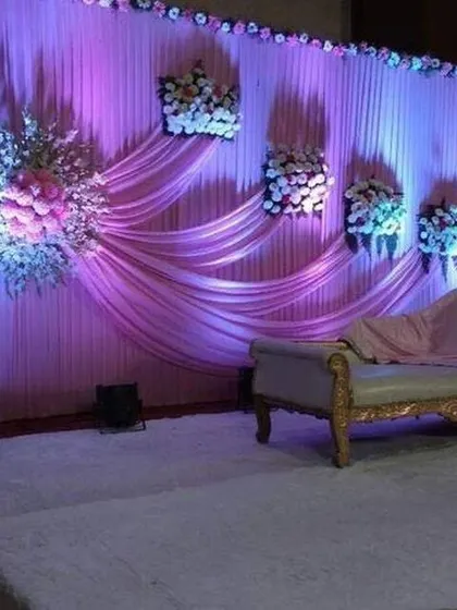 A simple yet elegant stage design perfect for an engagement or sangeet ceremony. The backdrop is made of soft, pleated purple fabric with strategically placed floral bouquets and uplighting to create a gentle, romantic mood.