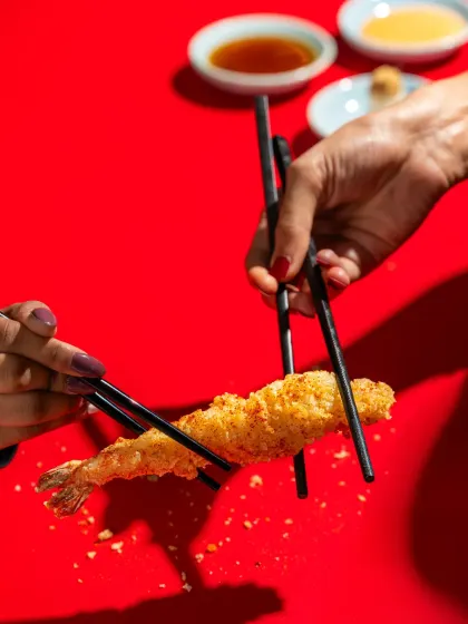 An action shot of two people using chopsticks to share a piece of shrimp tempura against a striking red background.