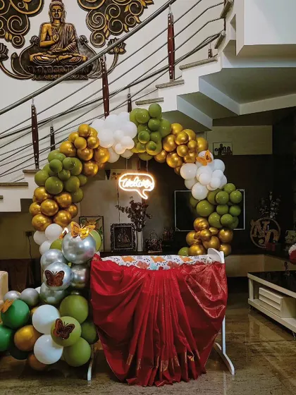 A beautiful and simple cradle decoration for a baby welcoming ceremony at home. The circular balloon arch in green, gold, and white, along with a 'Welcome' neon sign, creates a warm and inviting space.