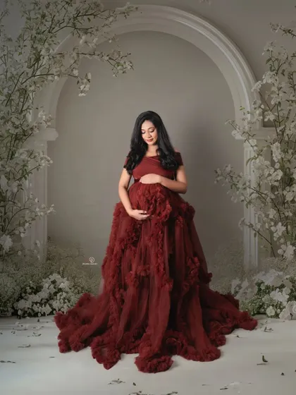 A moment of quiet reflection surrounded by a cascade of white flowers. This portrait in a deep red ruffled gown showcases the artistic and emotive potential of a studio maternity session.