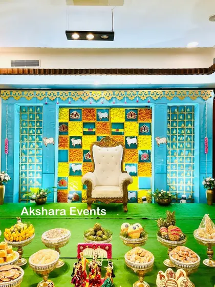 A vibrant Seemantham stage with a blue and yellow checkerboard backdrop. The decor includes traditional cow and lotus motifs, with a full display of offerings adding to the festive look.