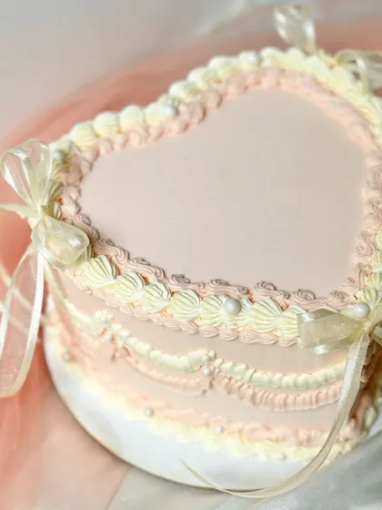 A close-up of a heart-shaped cake with delicate, multi-toned vintage piping. The soft pink, cream, and white ruffles create a beautiful, romantic texture.