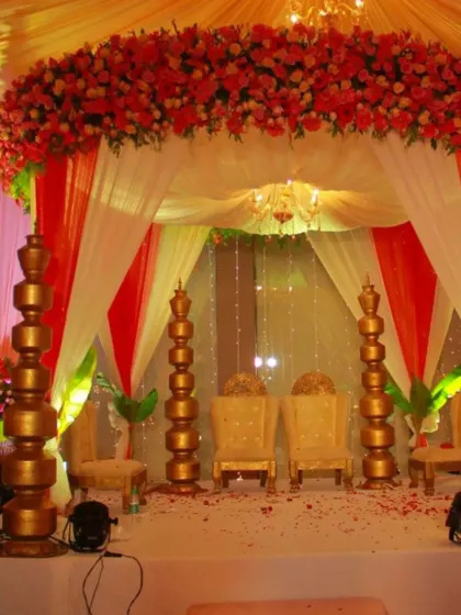 A classic wedding mandap design with vibrant red and white florals. The stage is set with traditional golden pillars and elegant seating for the bride and groom.