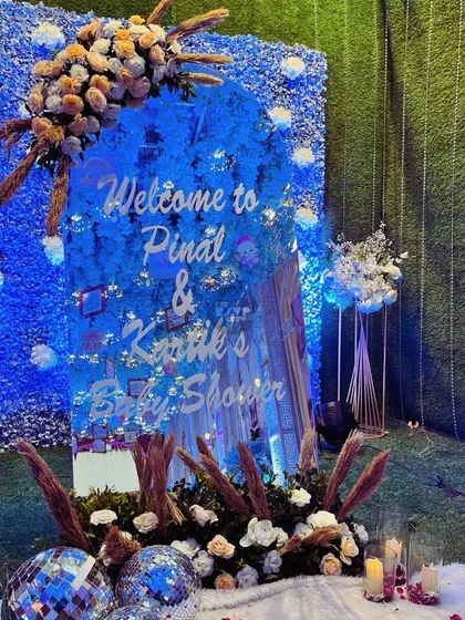 A custom acrylic welcome sign for a baby shower, set against a vibrant blue flower wall. The sign is adorned with pampas grass and floral arrangements, with disco balls at the base to add a touch of sparkle and fun.