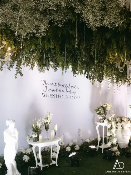 A romantic photo corner featuring a beautiful quote on a white wall, under a ceiling of hanging greenery and crystals. White statues and floral arrangements complete this dreamy setup.