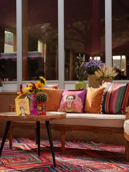 A seating area set against a large window, with the outdoor greenery visible. The decor features Frida Kahlo cushions and vibrant Birds of Paradise flowers.
