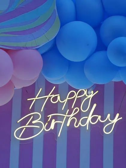 A close-up of the 'Happy Birthday' neon sign from a unicorn party. The pastel pink and blue striped backdrop adds to the playful and sweet theme.