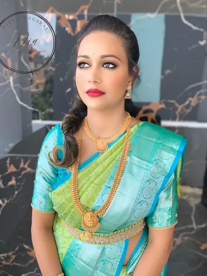 A subtle yet striking makeover for an engagement day. This look features a classic winged eyeliner and a bold red lip, which stands out beautifully against the bride's green and blue silk saree. It's a confident and gorgeous choice.