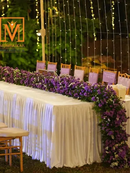 Elegant guest dining for a reception. A long table is decorated with a cascading runner of purple flowers, set under a canopy of fairy lights for a magical, intimate dinner experience.
