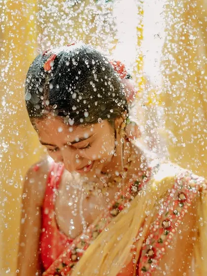 A close-up of the bride during the Haldi ritual, her face lit with joy as she is showered with water. The sunlight catches the water droplets, creating a beautiful, sparkling effect.
