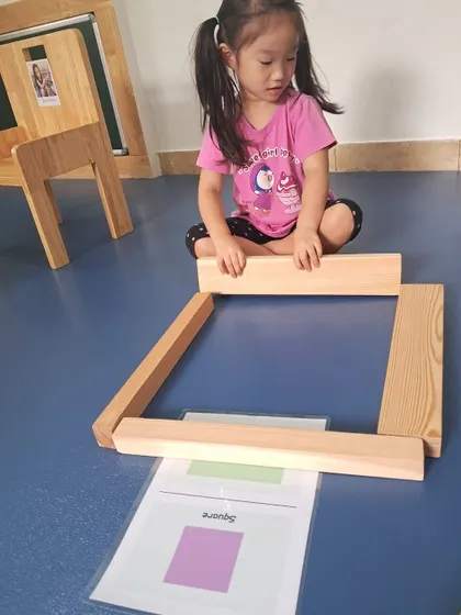 A child uses long wooden planks to build a large square on the floor, referencing a shape card. This activity connects abstract shapes with physical construction.